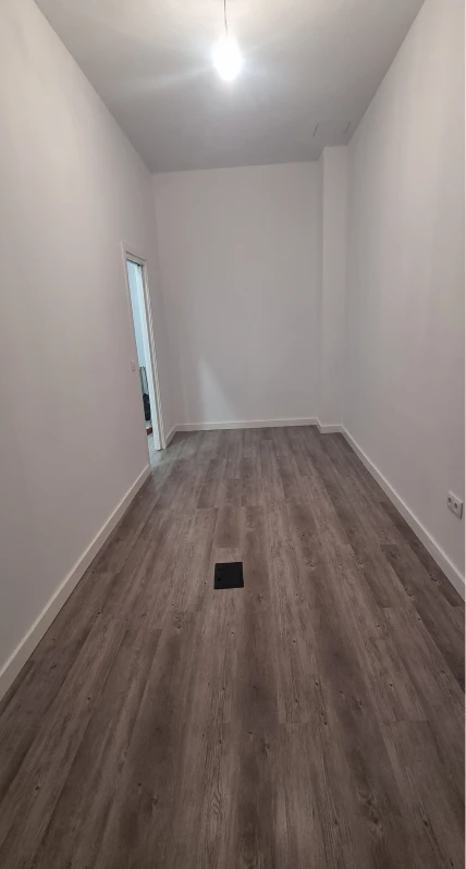 La imagen muestra una habitación con paredes blancas y un suelo de madera laminada de tono grisáceo. En el centro del suelo se observa una rejilla de ventilación. Hay una puerta a la izquierda, probablemente de acceso a otra área, y una lámpara de techo que proporciona luz en la habitación. El ambiente está desprovisto de muebles, lo que sugiere que aún se encuentra en proceso de acondicionamiento o es una estancia vacía.