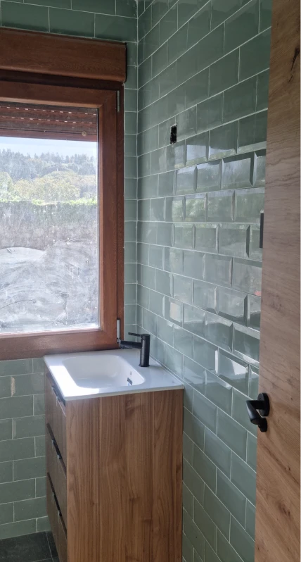 La imagen muestra un baño pequeño y moderno con azulejos de cerámica en color verde en las paredes. Hay un mueble de lavabo de madera con un diseño sencillo y elegante, con un grifo negro mate. La ventana permite que entre luz natural y ofrece una vista hacia el exterior. La puerta de madera está visible a la derecha de la imagen, y el ambiente tiene un estilo limpio y minimalista. El baño parece estar en proceso de acabados.
