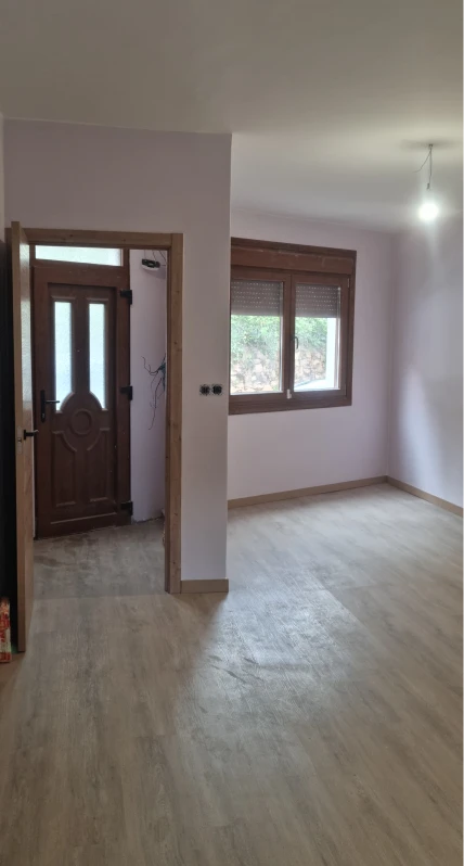 La imagen muestra una habitación recién terminada con un suelo de vinilo en tono madera clara, paredes pintadas de color lila y una ventana con persianas. La puerta de entrada, de madera, da paso a la habitación. El espacio tiene luz natural y aún no parece estar completamente amueblado, con solo un par de detalles visibles. El entorno parece ser de una casa o apartamento en proceso de ser amueblado.