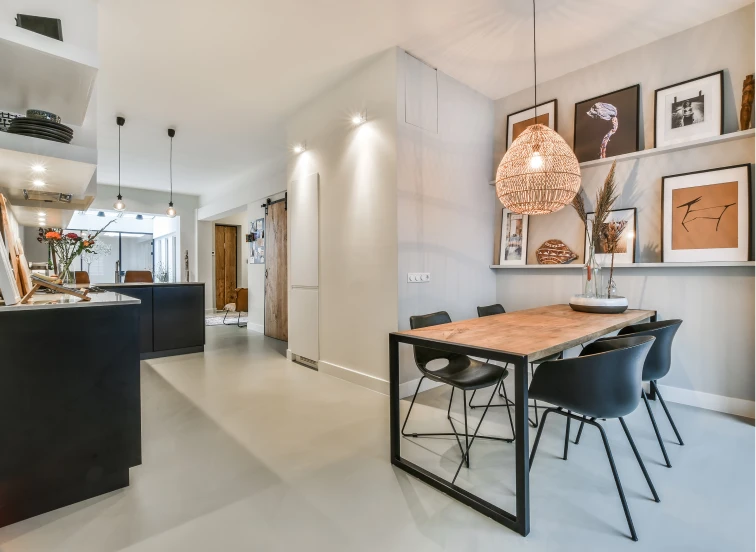 Comedor moderno y luminoso con una mesa de madera acompañada por sillas negras de diseño contemporáneo. La iluminación suave proviene de una lámpara colgante de estilo natural, mientras que las estanterías flotantes exhiben obras de arte y decoración, creando un ambiente acogedor y elegante para el hogar.